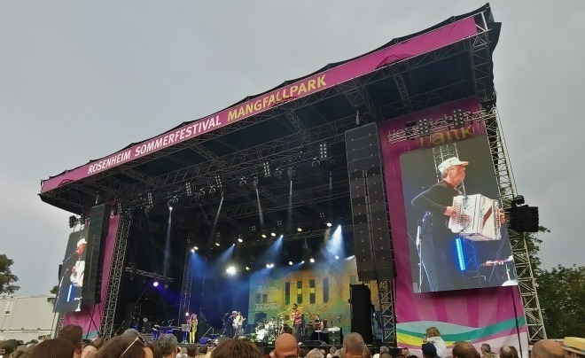 Foto: Hubert von Goisern steht mit einer Band auf der Bühne des Rosenheimer Sommerfestivals mit seiner Ziehharmonika und einer Ananas-Kappe auf dem Kopf.