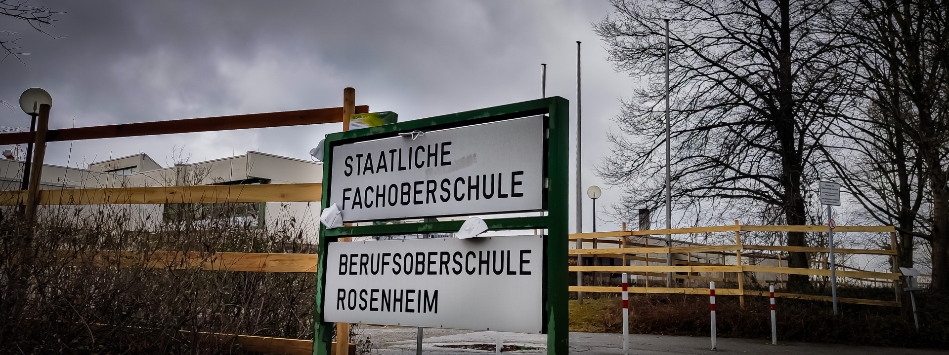 Foto eines Schildes mit der Aufschrift "Fachoberschule Rosenheim" und "Berufsoberschule Rosenheim", vor einem großen Gebäude. An dem Schild hängen verschiedene FFP2-Masken und auf dem Schild liegt eine Packung Schnelltests.