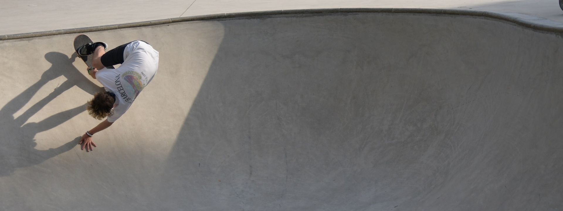 Das Foto zeigt einen Pool aus Beton zum Skaten. Links im Foto ist ein Skateboarder an der Wand des Pools und macht einen Trick. Die Sonne wirft seinen Schatten auf den Beton.