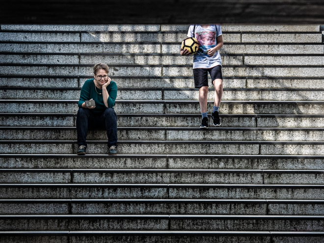 Foto: Auf einer großen Steintreppe sitzt eine freundlich blickende Frau, neben ihr ein junger Mensch mit Fußball, dessen Kopf nicht zu sehen ist