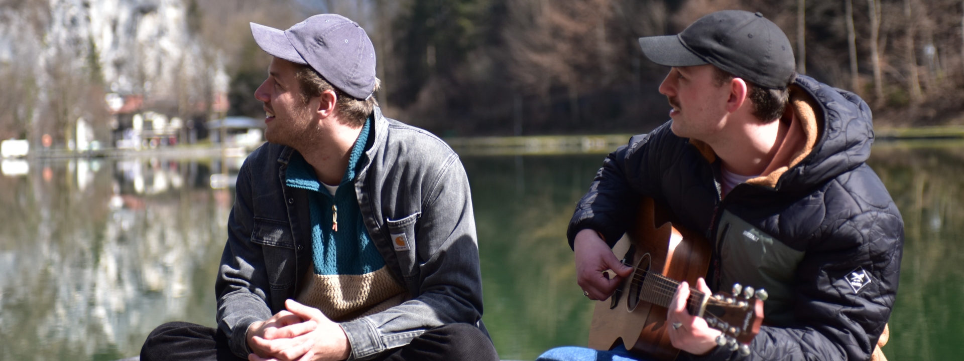 Foto: Zwei junge Männer (Gotti und Eisna), beide mit Basecap, sitzen im Schneidersitz auf einem Steg an einem See. Eisna spielt dabei Gitarre.