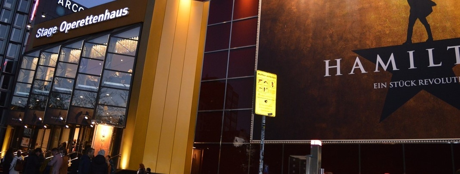 Foto: Das Stage Operetten Theater in Hamburg bei Nacht, gold-gelb beleuchtet mit einem Plakat und dem Schriftzug Hamilton und einem menschlichen Schattenriss