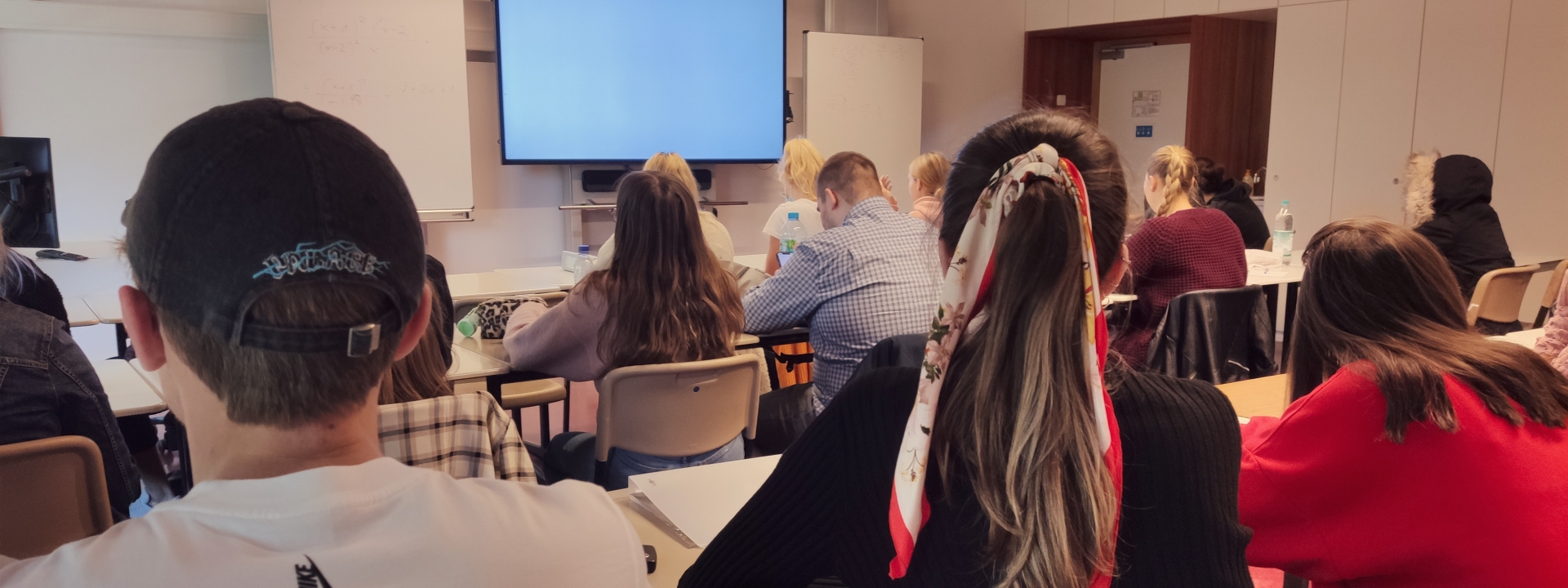 Foto eines Klassenzimmers, in dem Schüler direkt nebeneinander an Tischen sitzen, vorne ist eine große digitale Tafel