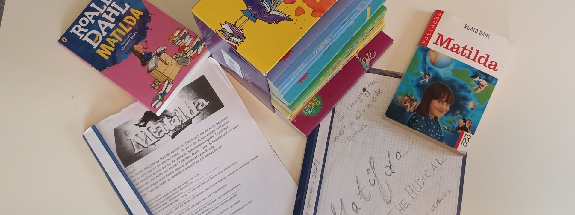 Von links nach rechts: Buch mit blau rosa cover Titel: "Matilda" darunter ein blauer Hefter mit dem Skript von Matilda, daneben ein gelber Buchschuber, daneben ein Buch auf dem Cover ein Bild von einem Mädchen vor blauem Hintergrund in Rotem Schriftzug da