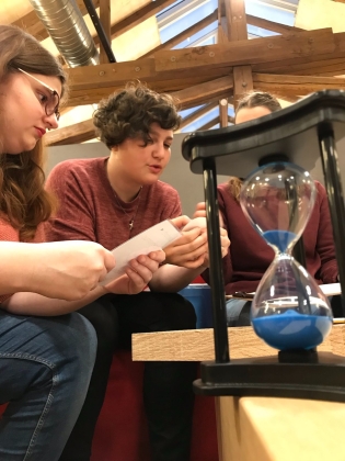 Foto: Zwei Jugendliche sitzen in einem Raum mit Holzbalken, im Vordergrund läuft eine große Sanduhr mit blauem Sand durch.