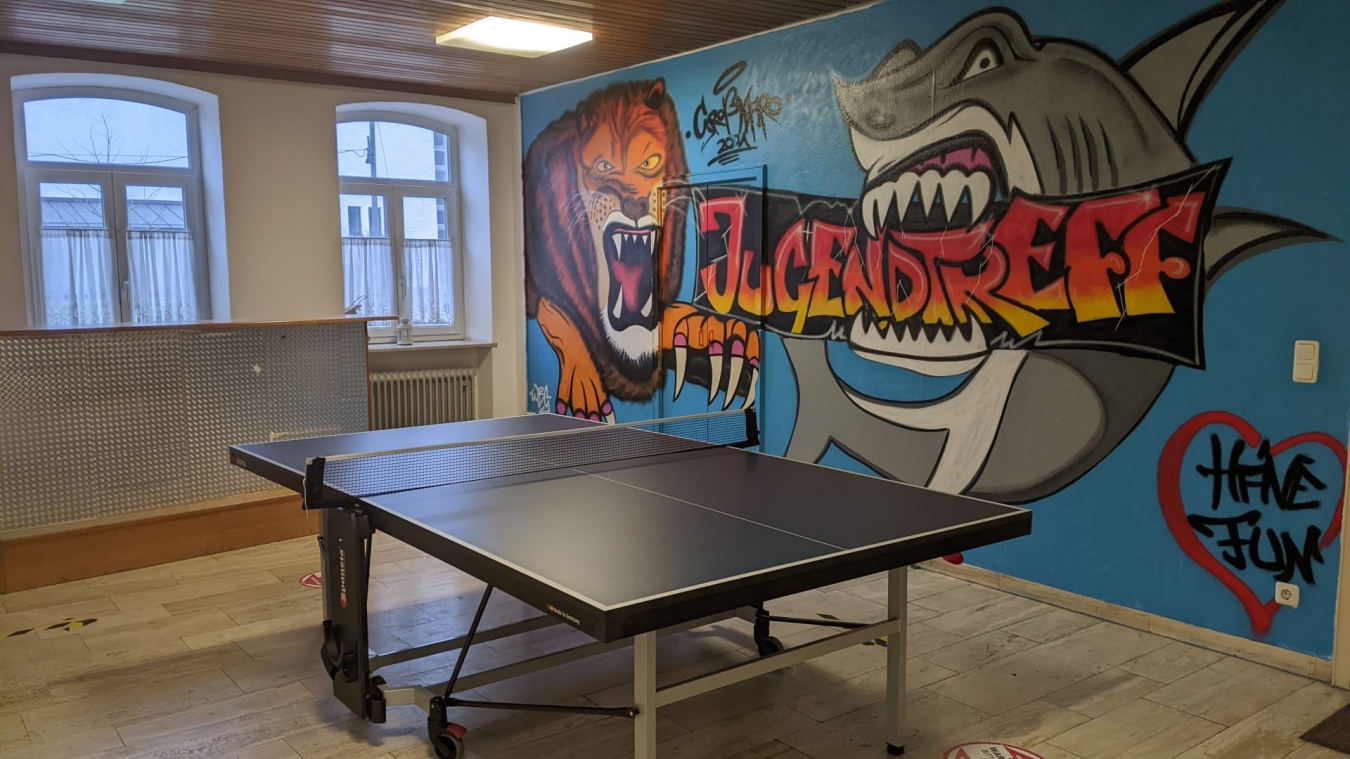 Foto: Eine Tischtennisplatte in einem Jugendraum. Im Hintergrund ein XXL Graffiti auf einer Wand mit dem Wort Jugendtreff, einem Hai und einem Löwen.
