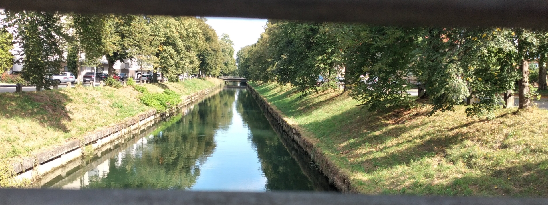 Bild des Mangfallkanals von einer Brücke herunter. Fotografiert zwischen zwei metallenen Gitterstäben hindurch. Man sieht rechts und links eine grüne Böschung, am Rand stehen ein paar Bäume.