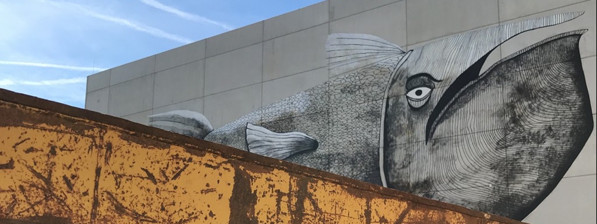 Foto: Im Vordergrund eine Ecke eines verrosteten Containers, dahinter ein Teil des großen Muralwalks, das einen Wal zeigt, der gleichzeitig ein Zeppelin ist.
