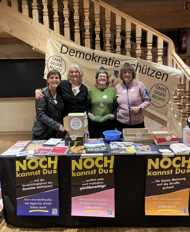Foto: 4 Frauen mit Buttons auf Pullovern und Jacken, an einem Aktionsstand vor einem Banner mit der Aufschrift 