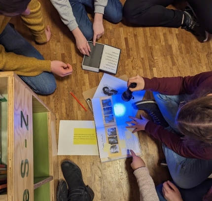 Beschäftigt beim Rätseln! | Foto: Jonas Ein Buch wird mit einer Schwarzlichttaschenlampe bescheint. Mehrere Jugendliche sitzen drum herum und zeigen drauf.