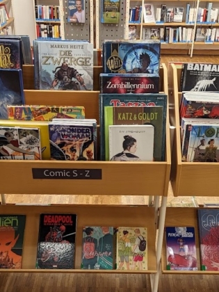 Foto: Ein Holz-Regal voll mit verschiedensten Comics. Im Hintergrund weitere Bücher.