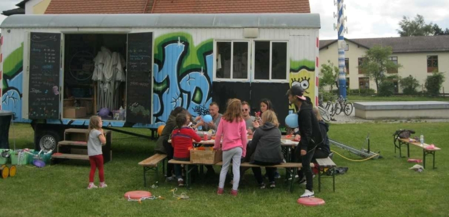 Foto: Ein bunt bemalter Bauwagen steht auf einer Wiese. Davor sitzen junge Betreuer mit Kindern an einem Biertisch