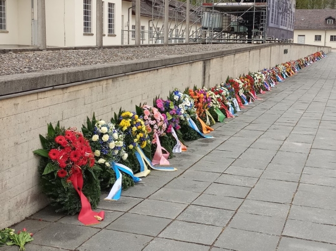 Foto: Viele bunte Blumenkränze an eine Mauer des KZs gelehnt