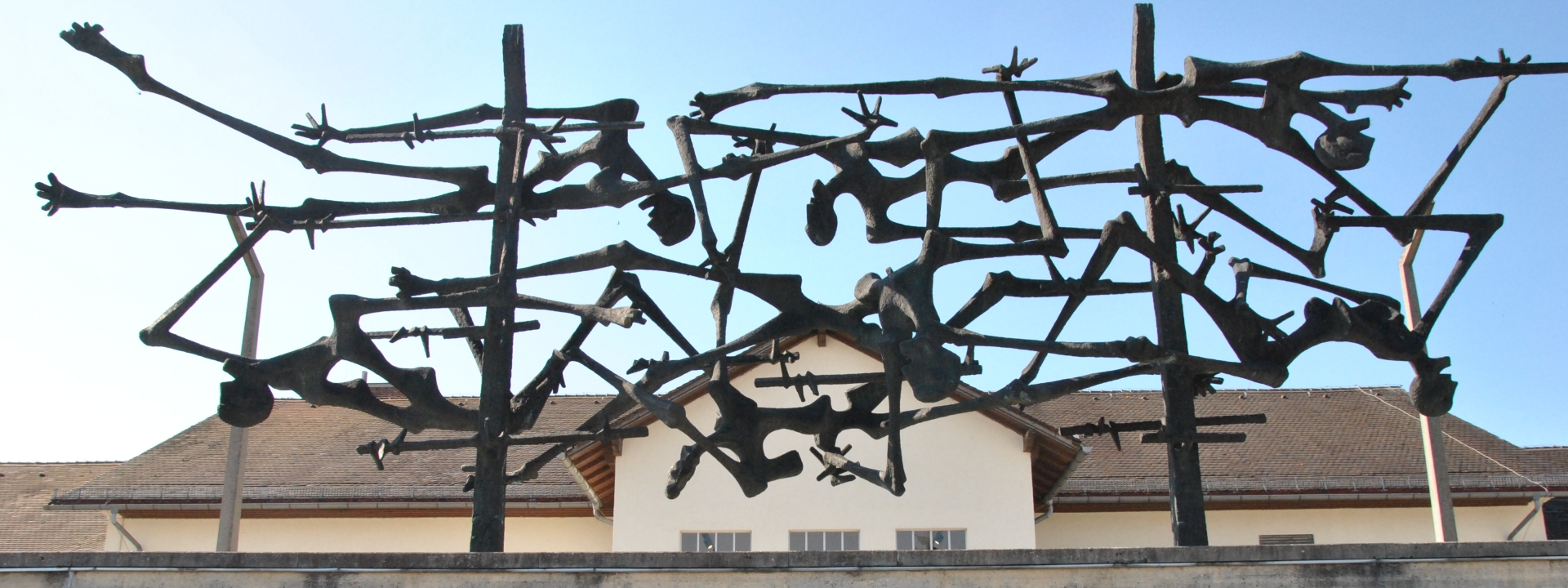 Foto: Ein Kunstwerk aus Eisen über einer Mauer stellt ausgemergelte menschliche Körper dar, die kreuz und quer durcheinander liegen