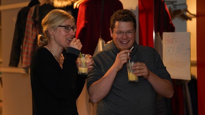 Foto: Zwei Erwachsene stehen nebeneinander, halten je ein Glas mit einem Coktail in der Hand und lächeln.