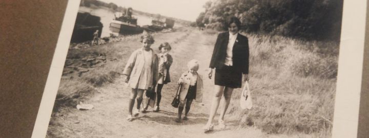 Schwarz-weiß-Foto aus einem Familienalbum: Drei Kinder und eine Frau gehen auf einem Schotterweg am Fluss entlang.