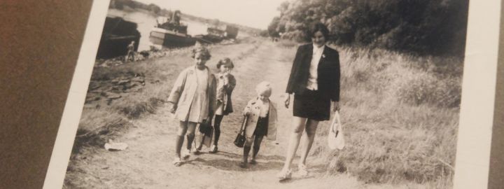 Aus dem Familienalbum: Erinnerungen an die DDR. Foto: Melina Schwarz-weiß-Foto aus einem Familienalbum: Drei Kinder und eine Frau gehen auf einem Schotterweg am Fluss entlang.