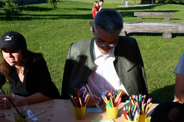 Foto: Landrat Otto Lederer sitzt neben einer Jugendlichen an einem Biertisch und notiert sich Fragen auf einem Zettel. Vor ihm stehen viele bunte Stifte in Bechern.