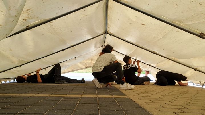 Foto: vier junge Menschen sitzen auf dem Boden unter einem weißen Zeltdach, das sie gerade aufbauen.