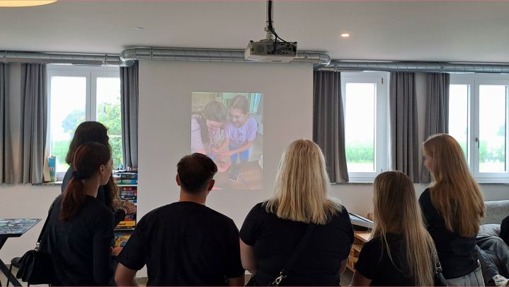 Foto: Fünf Junge Menschen stehen in einem Raum und schauen Fotos über einen Beamer auf einer Leinwand.
