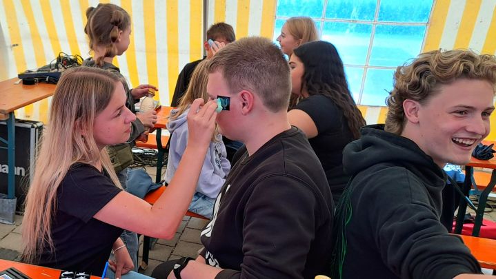 Foto: im Vordergrund sitzen drei junge Menschen auf einer Bierbank. Ein Junge lässt sich gerade von einem Mädchen ein kleines abwaschbares Tattoo ins Gesicht machen. Daneben sitzt ein Junge der sich freut.