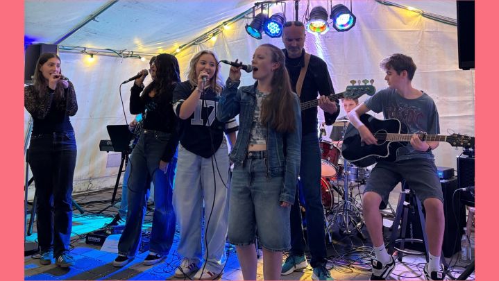 Foto: bei einem kleinen Konzert in einem bunt beleuchteten Zelt stehen im Vordergrund mehrere Sänger*innen am Mikrofon.im Hintergrund spielt ein Junge Gitarre.
