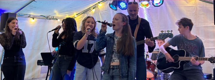 Foto: bei einem kleinen Konzert in einem bunt beleuchteten Zelt stehen im Vordergrund mehrere Sänger*innen am Mikrofon.im Hintergrund spielt ein Junge Gitarre, ein Junge Schlagzeug und ein Mann Bass.