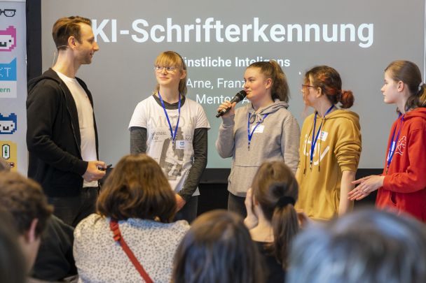 Foto: Ein Moderator stellt den vier Mädchen einer Gruppe bei ihrer Abschlusspräsentation zu KI-Schrifterkennung Fragen.