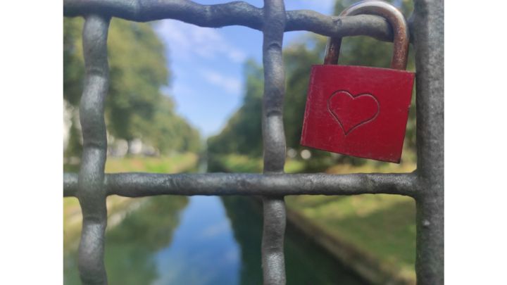 Foto: Scharf im Vordergrund ist ein Metallgitter, daran auf der rechten Seite ein rotes Liebesschloss mit Herz. Im Hintergrund der Mangfall-Kanal.