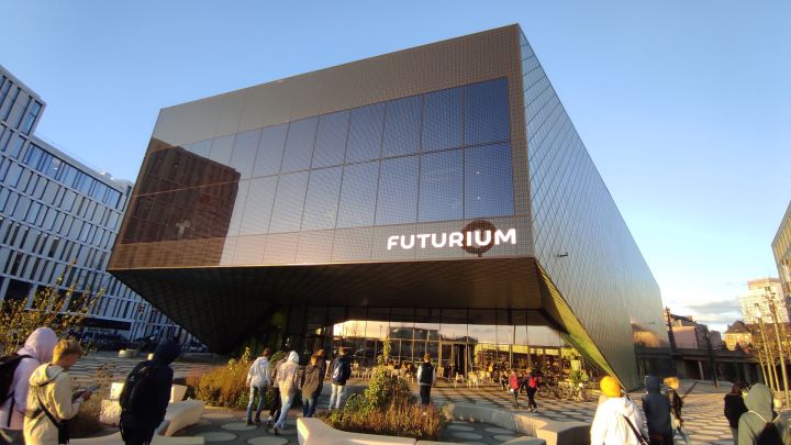 Ein braun-schwarzes, gläsernes Gebäude mit dem Namen FUTURIUM unten links. Es hat einen Vorsprung, somit ist der andere Gebäudeteil etwas weiter hinten. Links ist ein anderes Gebäude, rechts ist ein Teil eines Baumes. Oben ist des blaue Himmel
