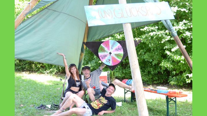 Foto: drei junge Menschen liegen unter einem Zelt in der Wiese, vor einem Glücksrad.