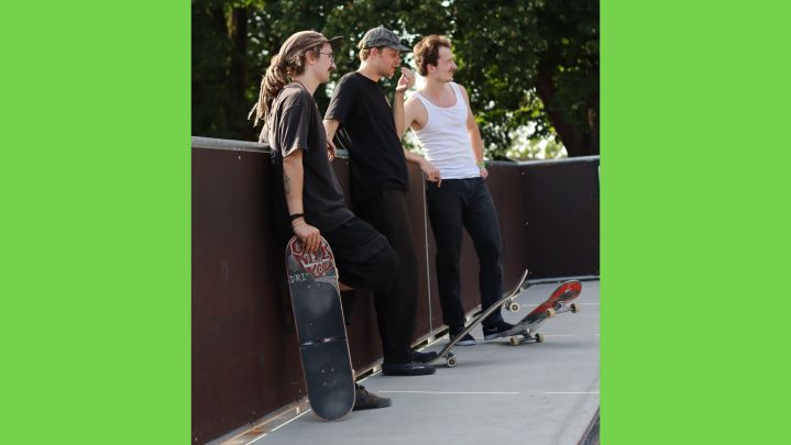 Foto: drei Skateboarder lehnen an einer Holzbande und halten ihre Skateboards in der Hand.