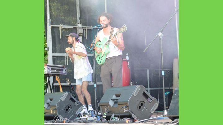 Foto: zwei Musiker auf einer Bühne. Einer spielt Gitarre und singt, der andere macht Percussion. Im Vordergrund sind Monitor-Boxen.