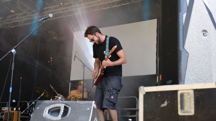 Foto: ein Musiker auf einer Bühne spielt Gitarre. Im Vordergrund sind Monitor-Boxen zu sehen. Im Hintergrund strahlt ein Scheinwerfer von unten  hinter dem Musiker hervor. 