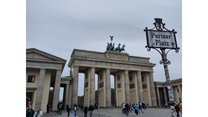 In der Mitte ist das beige Brandenburger Tor, ein Tor mit fünf Durchgängen, mit den grünen Figuren oben. Links ist ein Gebäude im selben Stil. Vorne rechts ist ein Schild mit der Aufschrift "Pariser Platz". Der Himmel ist bewölkt.