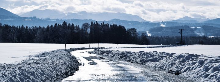 Auf dem Foto sieht man eine Straße im Winter. Es liegt sehr hoch Schnee und im Hintergrund sieht man die Berge. Die Farben sind Blautöne von fast weiß bis zu fast schwarz.