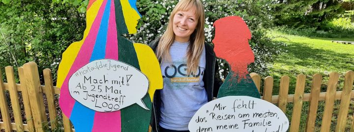 Foto: Eine Frau mit 2 bunt bemalten Holz-Figuren und Sprechblasen mit Aufschriften wie "Mir fehlt das Reisen am meisten, weil meine Familie im Ausland lebt"