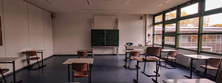 Foto: Menschenleerer Klassenraum mit Blick Richtung Tafel, mit einigem Abstand stehen Stühle vor Einzeltischen