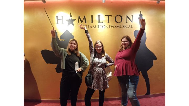 Foto: Drei junge Frauen vor einer gelb-goldenen Wand mit dem Titel "Hamilton", einen Arm erhoben und einen in die Hüfte gestützt.