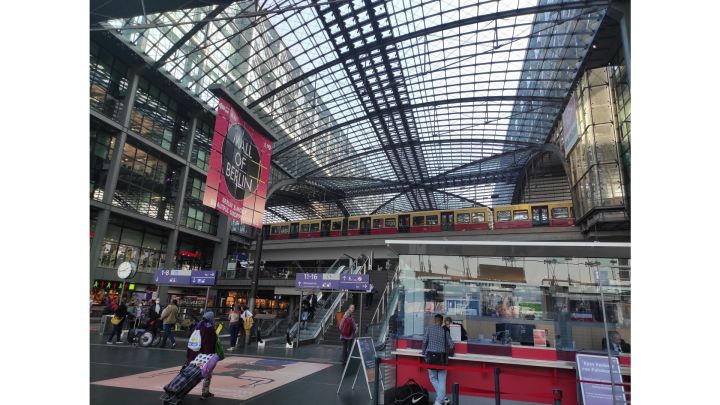 Der gläserne Hauptbahnhof. Unten sieht man den grauen Boden mit den Rolltreppen zu den unterirdischen Gleisen. In der Mitte fährt eine gelb-rote S-Bahn durchs Bild. Oben sieht man durch das Glas Dach den bewölkten Himmel.