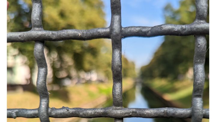 Foto: Im Vordergrund ist ein Brücken-Gitter, dahinter ein Kanal mit zwei Ufern und einigen Bäumen.