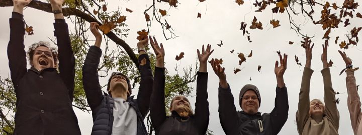 Foto: 5 junge Menschen werfen Herbstblaub in die Luft. Ihre Arme sind nach oben gestreckt.