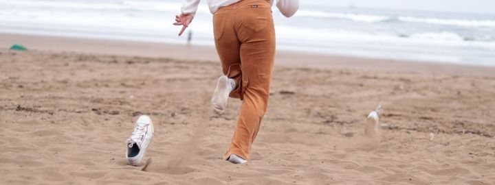 Foto: Eine Person am Strand von hinten, die auf Socken durch den Sand auf das Meer zuläuft. Die Sneaker, die sie gerade abgestreift hat, fliegen durch die Luft.