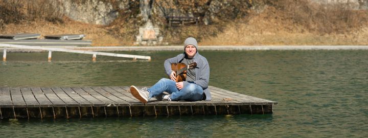 Foto: Ein junger Mann mit Mütze und Gitarre sitzt auf einem Holzsteg an einem See