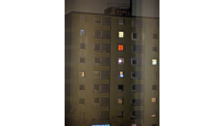 Foto: Blick durch ein geschlossenes Fenster auf einen Wohnblock. In manchen Fenstern scheint Licht, in einem ein großer Stern.
