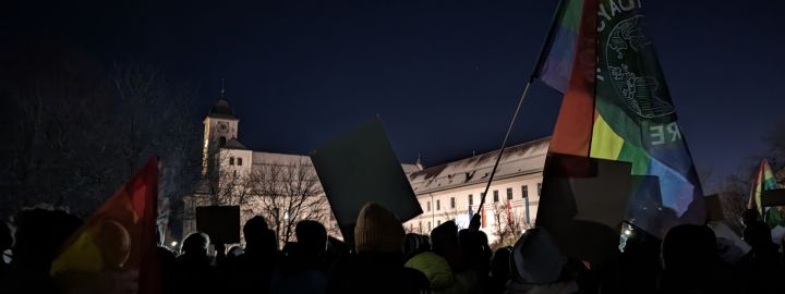 Foto: Die Rücken von vielen Menschen, die am dunklen Abend vor dem beleuchteten Kloster in Rott mit Plakaten und einer Regenbogenfahne demonstrieren