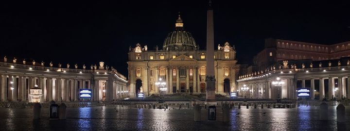 Foto: Der Petersplatz in der Vatikanstadt bei Nacht. Die Gebäude sind beleuchtet, der Platz ist menschenleer, das Kopfsteinpflaster schimmert nass.
