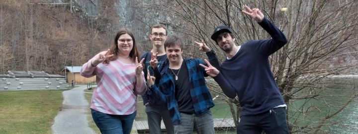 Foto: Vier Personen, zwei davon mit Down-Syndrom, lachen in die Kamera und zeigen mit den Fingern Victory Zeichen. Im Hintergrund ein See.