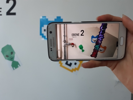Foto: Ein Smartphone filmt eine Stellwand mit Stickern. Auf dem Bildschirm sieht es so aus, als würde auch noch ein Graffiti auf der Wand kleben.
